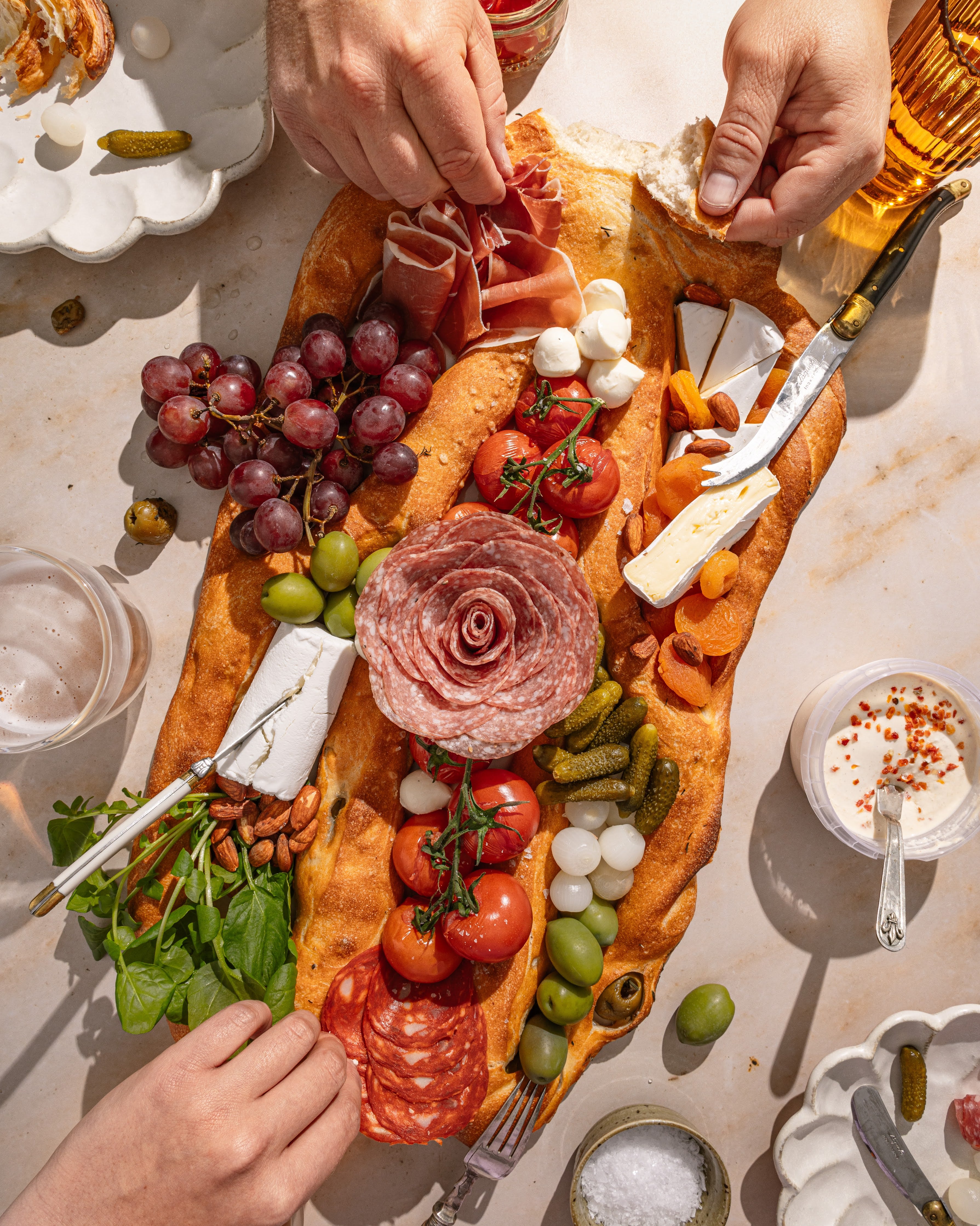 A fougasse hosting platter with salami, olives, tomatoes, cheese and grapes, being pulled apart by two sets of hands